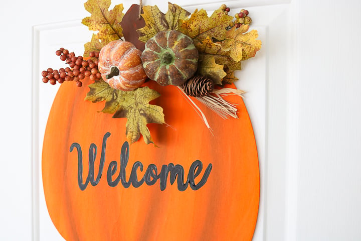 Painted wooden pumpkin door hanger with fall trim and Welcome lettering on the pumpkin