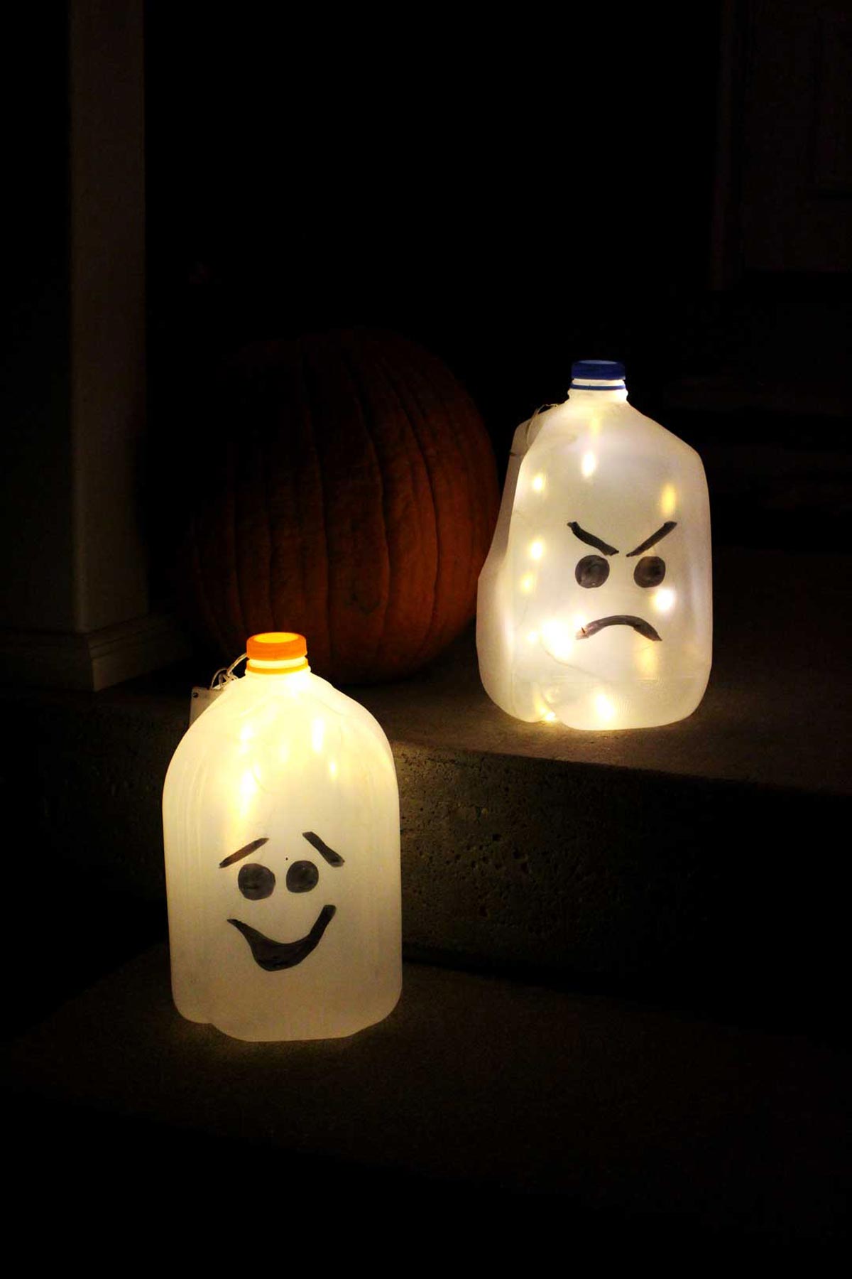 Two recycled milk jug ghosts with painted faces glowing on porch steps beside a pumpkin at night